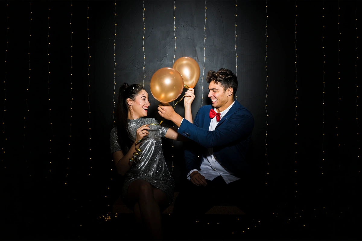 A couple in evening wear with gold and black balloons before a sparkling curtain