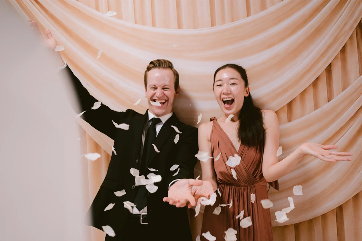 A couple laughing as white petals fall in front of draped champagne fabric